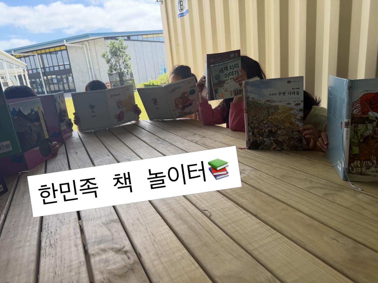 야외 테이블에서 함께 책을 읽는 한민족 책 놀이터 학생들